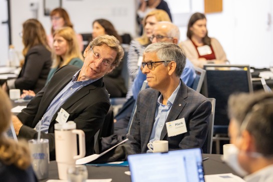 Attendee at summit leans over to converse with another attendee seated at a nearby table.